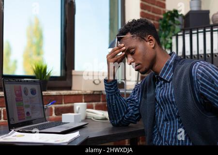 Tired marketing specialist feeling depressed because of startup low profit analytics on laptop display. African american office worker having a headache sitting at desk working with business charts. Stock Photo
