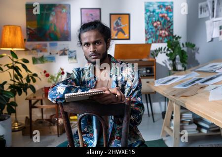 Self-confident young man of Indian ethnicity in smart casualwear working in art studio while sitting against walls with paintings Stock Photo