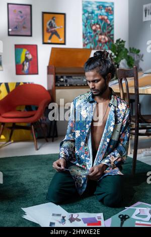 Creative guy in fashionable shirt selecting photos for creative collages while sitting on the floor in living room or art studio Stock Photo