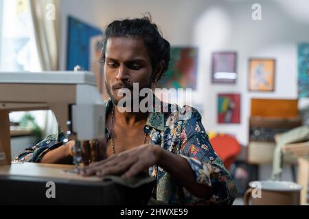 Serious stylish male tailor in non-gendered clothing creating new apparel while sitting in front of camera by electric sewing machine Stock Photo
