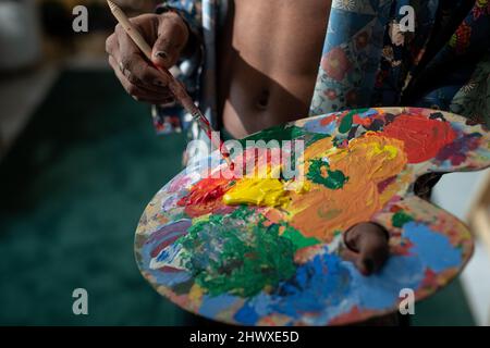 Hands of young non-binary man with paintbrush mixing acrylic paints on palette while creating new masterpiece in art studio Stock Photo