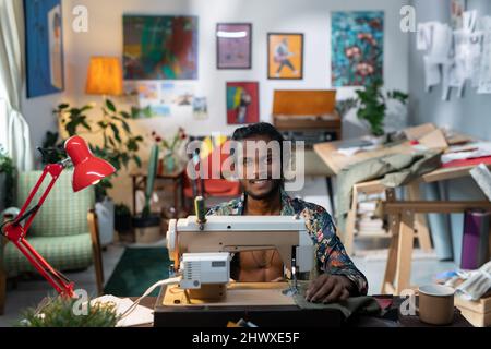 Young successful gender-neutral male designer of fashionable apparel working by sewing machine while sitting in workshop Stock Photo