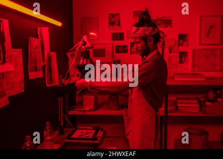 Young serious male photographer using photo enlarger while making black and white photographs in red lit laboratory Stock Photo
