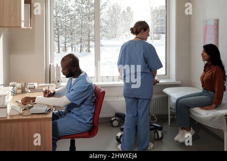 Young female patient waiting for ultrasound examination and talking to nurse while male doctor making notes or prescriptions Stock Photo