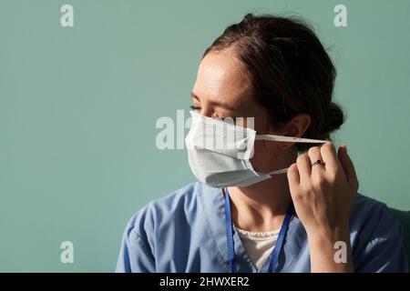 Young female doctor in blue uniform going to take off protective mask after tiresome work with sick patients in hospital Stock Photo
