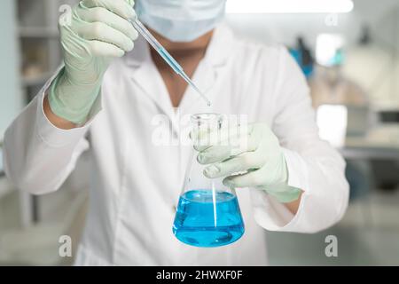 Contemporary scientist in surgical gloves, mask and lab coat adding drop of liquid into flask with blue chemical substance Stock Photo