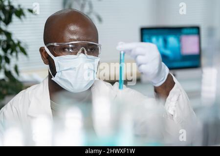 Young black man in lab coat, protective mask and gloves looking at flask containing blue liquid during work in laboratory Stock Photo