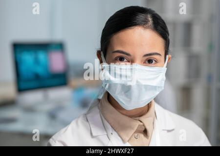 Young pretty female clinician in whitecoat and protective mask on face looking at camera while standing in medical office Stock Photo