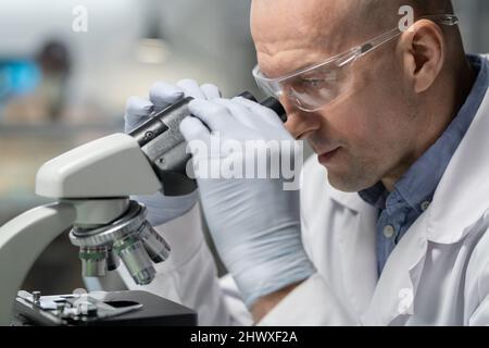 Gloved mature male scientist or chemist in lab coat researching chemical elements in microscope during investigation Stock Photo