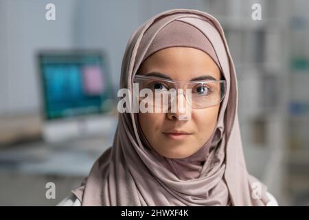 Face of young pretty Muslim female clinician in hijab and protective eyewear looking at camera against her workplace Stock Photo