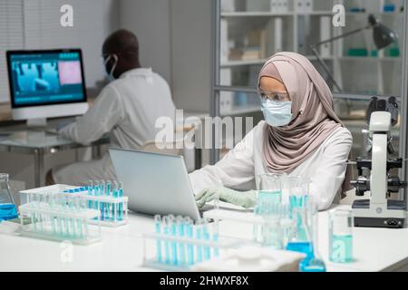 Muslim female scientist in hijab and protective workwear typing on laptop keyboard while sitting by workplace against coworker Stock Photo