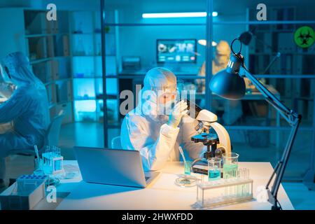 Female scientist in biohazard suit, respirator and gloves using microscope for studying new substances and their characteristics Stock Photo