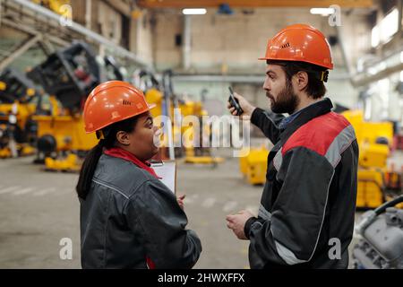 Two young confident interracial engineers in workwear having discussion of working points while standing in distribution warehouse Stock Photo
