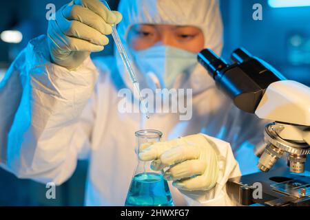 Gloved hands of contemporary chemist in coveralls and respirator experimenting with new chemical substances Stock Photo