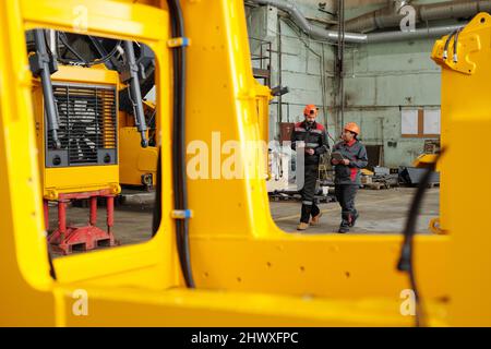 Two young engineers in helmets and workwear having discussion while moving along workshop of large modern industrial plant Stock Photo