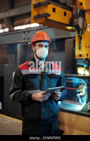 Young serious engineer in respirator, hardhat and workwear searching online technical data while using digital tablet by workplace Stock Photo