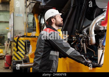 Young technician or repairman in hardhat and uniform standing by industrial machine while checking its engine system Stock Photo