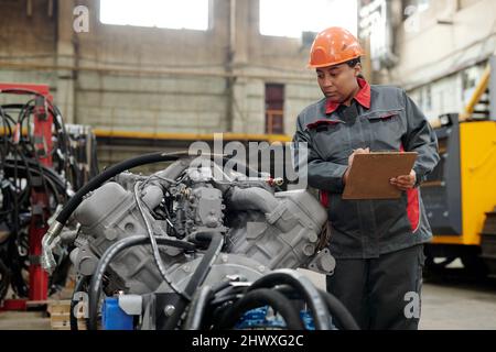 Young serious African American female mechanic making notes in document while checking motor of industrial machine Stock Photo