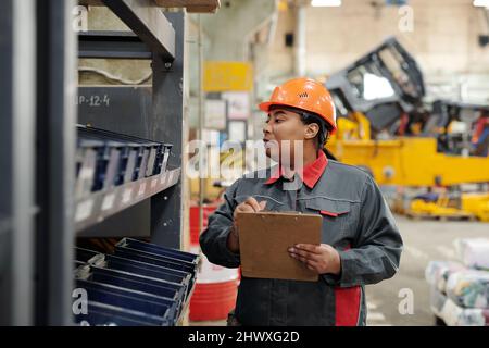 Female worker of industrial plant making revision notes in document while looking through several plastic containers with spare parts Stock Photo