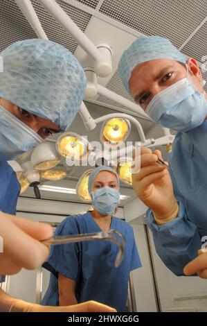 A surgical team of three doctors – two men and one woman – wearing blue gowns, caps and masks, inside a hospital operating theatre underneath overhead Stock Photo