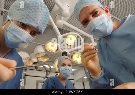 A surgical team of three doctors – two men and one woman – wearing blue gowns, caps and masks, inside a hospital operating theatre underneath overhead Stock Photo