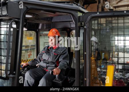 Constructor in hardhat and workwear sitting in cab of construction machine and driving it while keeping hands on gearshifts Stock Photo