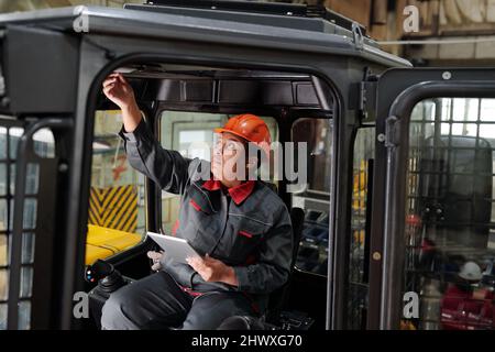 African American female worker of industrial plant with tablet driving construction machine while testing new vehicle Stock Photo