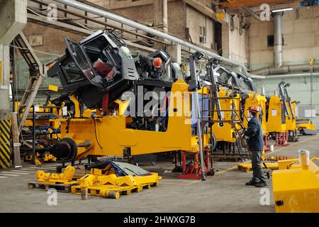 Two male workers of modern industrial plant or repairmen checking efficiency of huge construction machines in workshop Stock Photo