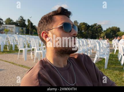 A portrait of handsome man with sunglasses in summer season. Close up photo. Empty chairs behind him. He is watching screen. Stock Photo
