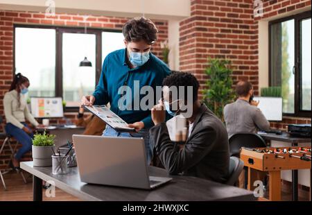 Diverse entrepreneurs working at company collaboration analyzing marketing graph discussing business ideas in brick wall startup office. Multi-ethnic team wearing medical face mask against covid19 Stock Photo