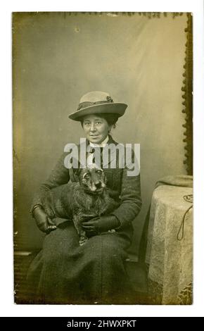 Original post Edwardian era postcard of middle-aged woman wearing fashions of the time - a two-piece tweed suit, long skirt, hat and gloves, with an old wire-haired corgi (possibly) sitting on her lap. Dated 1911, U.K. Stock Photo
