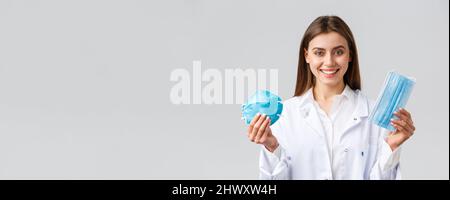 Covid-19, preventing virus, healthcare workers concept. Smiling attractive female doctor in white scrubs showing two variants of personal protective Stock Photo