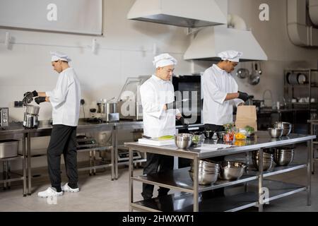 Multiracial team of cooks actively work in the kitchen. Asian chef announces order list from the printed check, Latin guy packs food for deliver, European cooks cooking behind. Concept of teamwork at restaurant Stock Photo