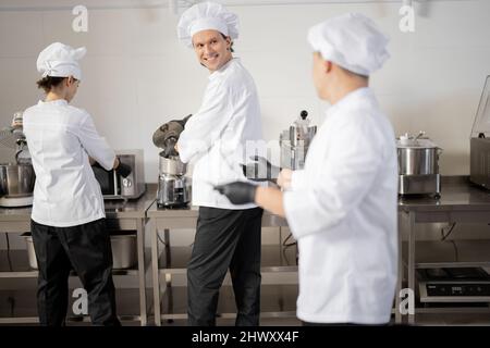 Multiracial well-dressed cooks cheering each other while cooking in professional kitchen. Concept of teamwork at restaurant Stock Photo