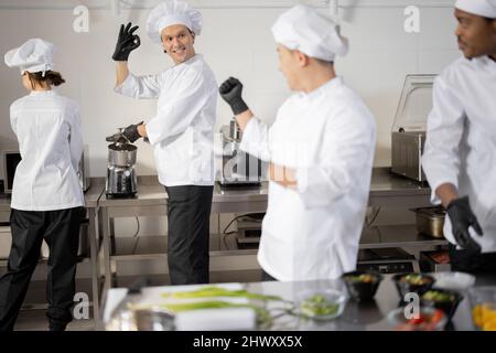 Multiracial well-dressed cooks cheering each other while cooking in professional kitchen. Concept of teamwork at restaurant Stock Photo