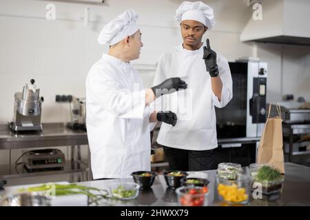 Asian and Latin chefs reading printed order while cooking meals in professional kitchen. Two chefs of different nationalities cook together in a restaurant Stock Photo
