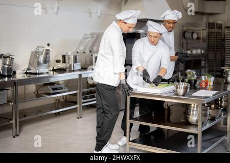 Three multiracial chefs having some conversation while cooking in professional kitchen. Interior with professional equipment and stainless tables. Teamwork at restaurant Stock Photo