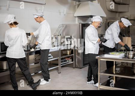 Happy multiracial team of cooks actively work in the kitchen. Asian chef announces order list from the printed check, Latin guy packs food for deliver, European cooks cooking behind. Concept of teamwork at restaurant Stock Photo