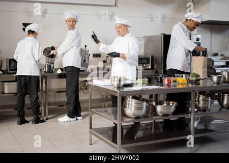 Multiracial team of cooks actively work in the kitchen. Asian chef announces order list from the printed check, Latin guy packs food for deliver, European cooks cooking behind. Concept of teamwork at restaurant Stock Photo