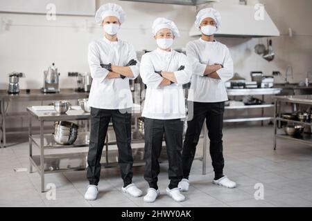 Full body portrait of multiracial team of three chefs standing together in the professional kitchen. Well-dressed chefs in face masks and protective gloves ready for a job. New normal for business during pandemic Stock Photo