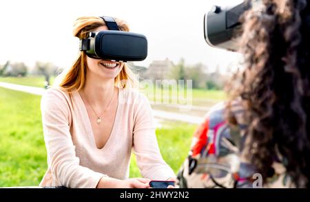 Young women couple playing on vr outdoors - Virtual reality and wearable tech concept with happy girls having fun together with headset goggles - Gene Stock Photo