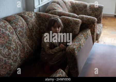 little Ukrainian girl is hiding in the sofa, looks confused Stock Photo