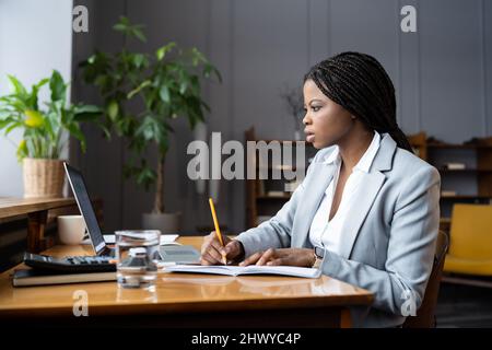 Concentrated young african businesswoman watching online business webinar and writing down notes Stock Photo