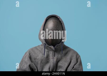 an unrecognizable man in a gray hoodie with a mask on his face isolated on a blue background, copy space Stock Photo