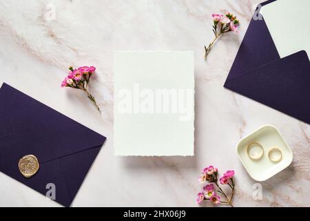 Wedding invitation card template, purple envelopes, rings, pink flowers on stone desk table. Flat lay, top view, copy space. Stock Photo