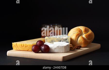 On the board are three types of cheese, next to a jar of honey and buns, nuts and grapes Stock Photo