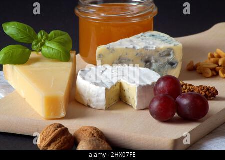 Three types of cheese, a jar of delicious honey, nuts, grapes and basil are stacked on a wooden board. The photo was taken against a black background. Stock Photo