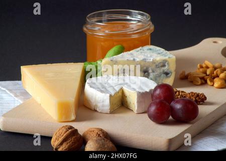 Three types of cheese, a jar of delicious honey, nuts, grapes and basil are stacked on a wooden board. The photo was taken against a black background. Stock Photo