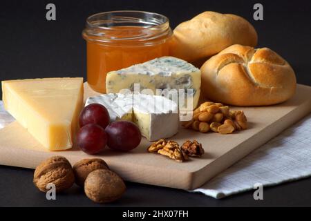 Three types of cheese, a jar of delicious honey, nuts, grapes and basil are stacked on a wooden board. The photo was taken against a black background. Stock Photo