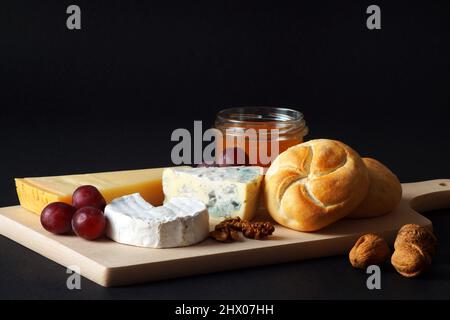 Three types of cheeses with honey, nuts, grapes and fresh delicious soft buns are shot on a black background. Excellent breakfast Stock Photo